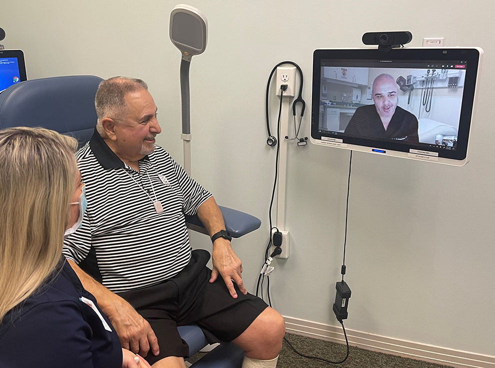 Dr. Bob Rahmanian, Cypress at Home medical director is pictured on-screen. Tisha Saylor, Cypress at Home manager of medical care services facilitates a telehealth visit with a Cypress Cove resident in the on-campus clinic, exam room 2.